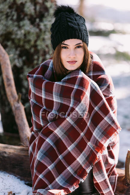 Zufriedene, in warmes Plaid gehüllte Frau steht in verschneiten Wäldern und blickt in die Kamera — Stockfoto