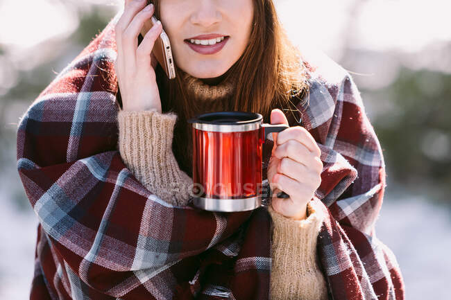 Cheerful female wrapped in warm plaid sitting on tree trunk in winter forest and talking mobile phone on sunny day — Stock Photo