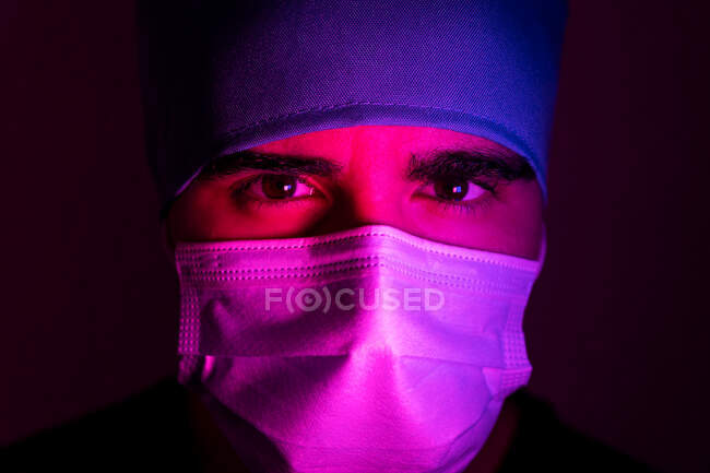 Primer plano del cirujano masculino en máscara médica mirando a la cámara en habitación oscura con luz de neón azul y roja - foto de stock