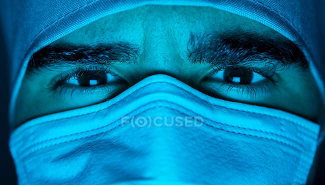 Primo piano del chirurgo maschio in maschera medica guardando la fotocamera nella stanza buia con luce blu al neon — Foto stock