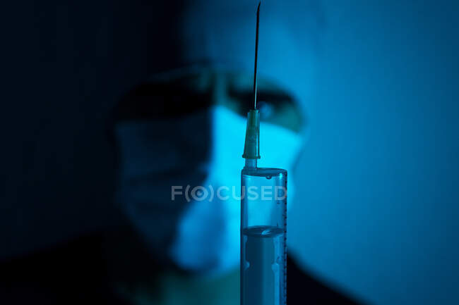 Анонимный хирург в маске со шприцем, наполненным химической жидкостью в темной комнате с голубым неоновым светом — стоковое фото