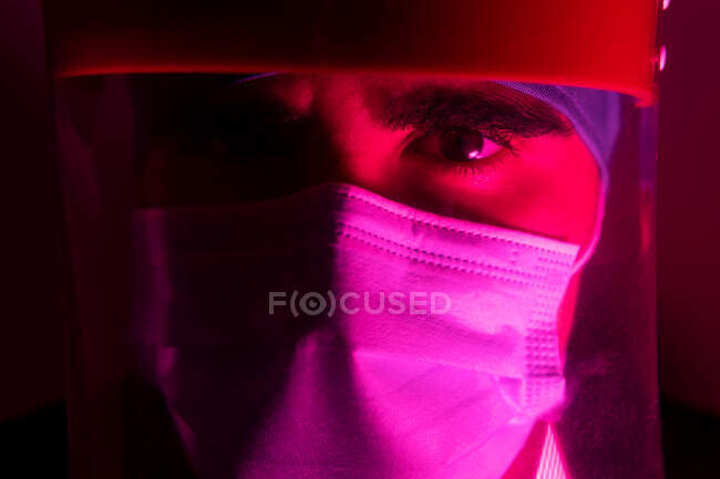 Gros plan du chirurgien masculin dans un masque médical regardant la caméra dans une pièce sombre avec une lumière au néon rouge — Photo de stock