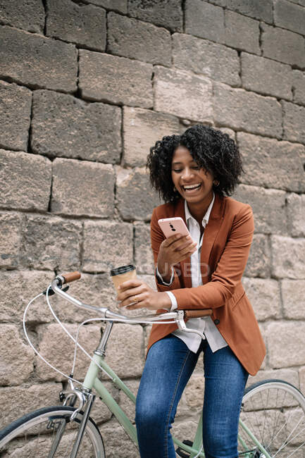 Encantada mujer afroamericana parada cerca de la bicicleta estacionada con bebida para llevar y mensajería en las redes sociales en el teléfono inteligente en la calle - foto de stock