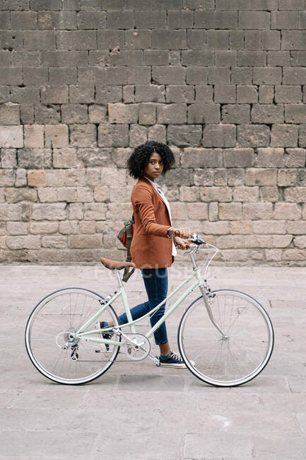 Vue latérale de la femme noire dans un style décontracté intelligent marchant le long de la rue avec vélo et regardant la caméra — Photo de stock