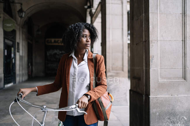 Black woman in smart casual style walking along street with bike and looking away — Stock Photo