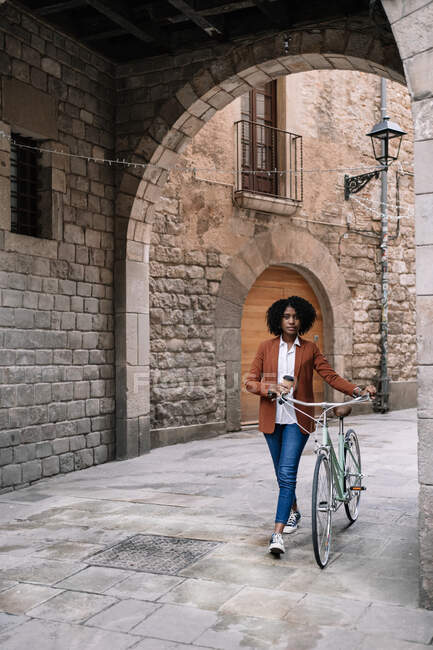 Mujer negra en estilo casual inteligente caminando por la calle con bicicleta y mirando hacia otro lado - foto de stock
