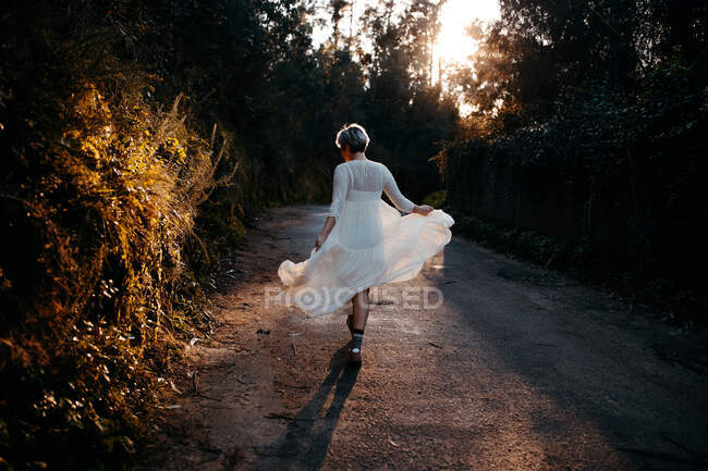 Full body back view of anonymous female wearing white dress walking on rural road among green trees in nature on evening time - foto de stock