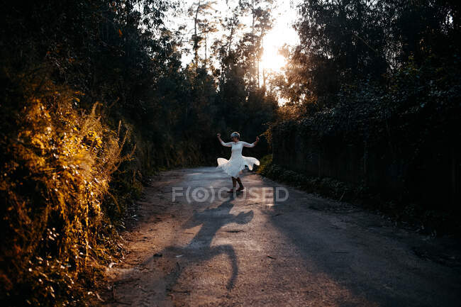 Full body of anonymous female wearing white dress walking on rural road among green trees in nature on evening time — Foto stock