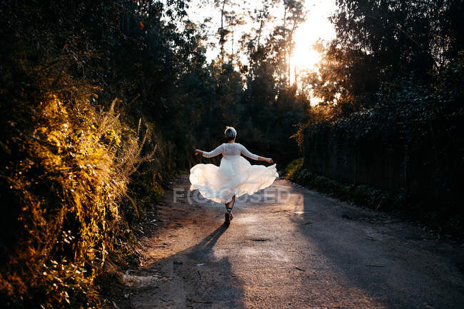 Full body back view of anonymous female wearing white dress walking on rural road among green trees in nature on evening time - foto de stock