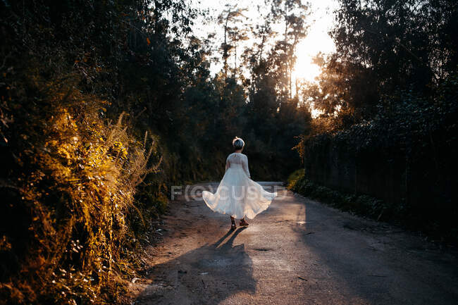 Vue arrière du corps complet de la femelle anonyme portant une robe blanche marchant sur la route rurale parmi les arbres verts dans la nature le soir — Photo de stock