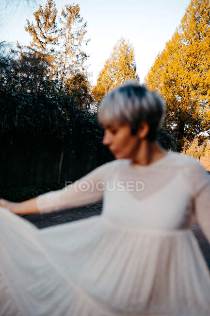 Side view of blurred anonymous female with short hair wearing white dress standing against tall green trees in rural terrain — Fotografia de Stock