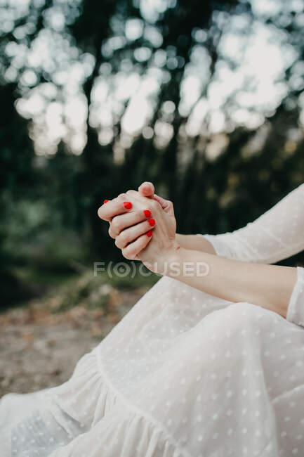 Unrecognizable female with red manicure wearing long white dress sitting on ground in forest with green trees on blurred background — Foto stock