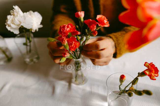 Alto ángulo de cultivo florista masculino anónimo sentado a la mesa con claveles y tulipanes en cristalería - foto de stock