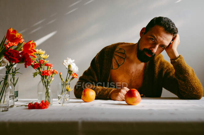 Der bärtige, nachdenkliche Mann lehnt an einem Tisch mit reifen Äpfeln und Blumen in Gläsern — Stockfoto