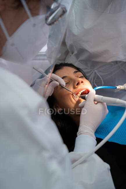 Профессиональный стоматолог в форме с медицинской маской сверлит зуб спокойной женщины с помощью ассистента — стоковое фото