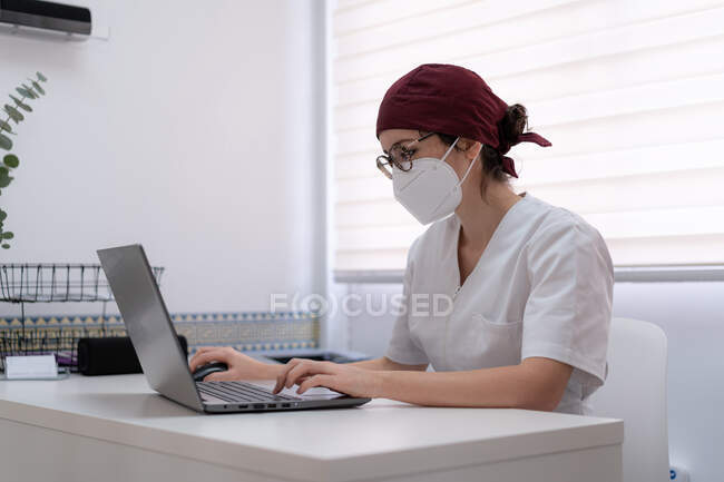 Konzentrierte Ärztin in medizinischer Maske und Uniform sitzt am Tisch und blättert im Laptop, während sie in modernen Arbeitsräumen arbeitet — Stockfoto