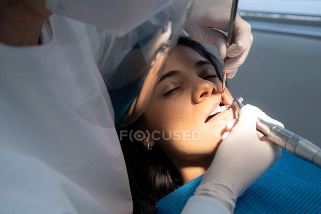 Профессиональный стоматолог в форме с медицинской маской сверлит зуб спокойной женщины с помощью ассистента — стоковое фото