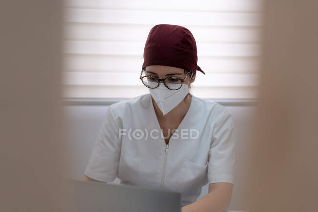 Концентрована жінка-лікар в медичній масці та уніформі сидить за столом та переглядає ноутбук під час роботи в сучасному робочому просторі — стокове фото