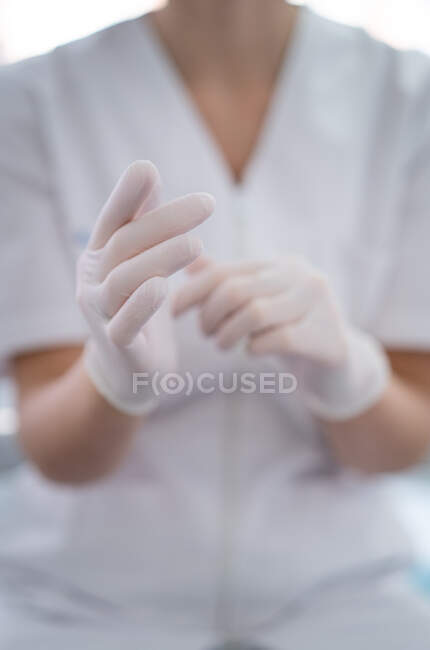 Crop unrecognizable female doctor in white uniform putting on clean latex gloves while working - foto de stock