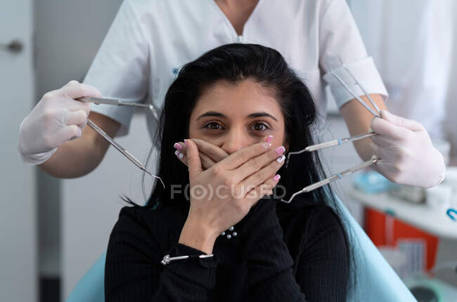 Jeune patiente effrayée regardant la caméra et couvrant la bouche des mains avec un médecin des cultures tenant des outils dentaires stériles — Photo de stock