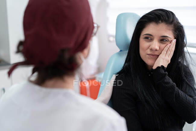 Back view of unrecognizable doctor and displeased female patient in casual clothes touching cheek while sitting in light workplace — Fotografia de Stock