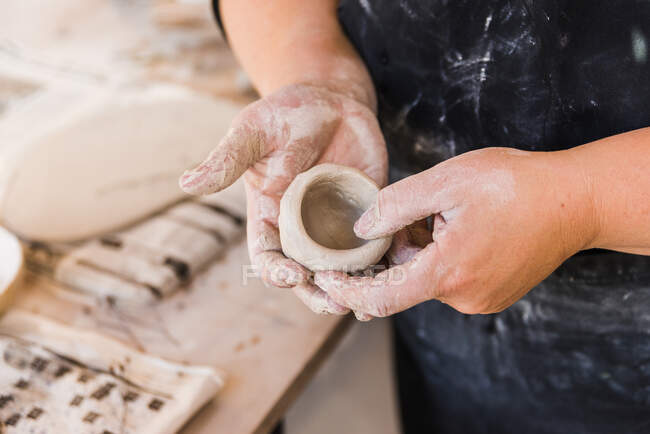 High angle of crop anonymous master in black dirty apron standing in workshop and shaping piece of clay in hands near table with tools for handicraft — Foto stock
