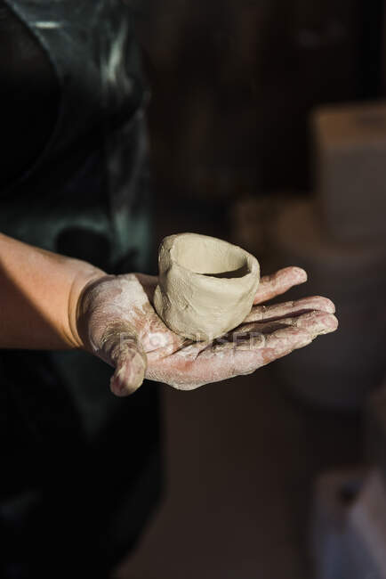 Cropped unrecognizable adult female artisan standing in light studio and holding fresh handmade ceramic pot - foto de stock