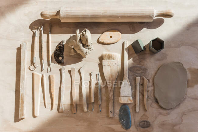 Верхний вид набора различных инструментов для керамики с петлями и стеллажами с кистями и скалкой с кусочком гладкой глины, размещенной на деревянном столе в мастерской — стоковое фото