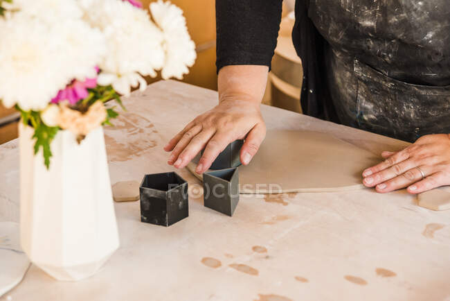 Corte fêmea irreconhecível em avental e roupa casual preta usando objetos geométricos para cortar formas em argila na mesa perto do vaso com flores na oficina — Fotografia de Stock