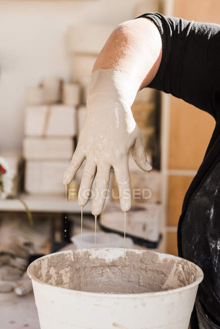 Анонимная ремесленница в грязной черной одежде, стоящая у стола и вытаскивающая грязную руку из ведра, наполненного глиной в студии при дневном свете — стоковое фото