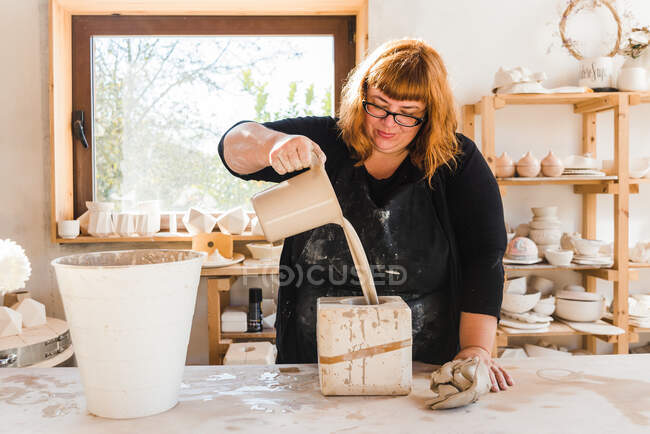 Adult craftswoman in black apron and casual clothes standing near table and pouring clay into a mold near bucket and window near shelves with earthenware in workshop in daytime — Fotografia de Stock