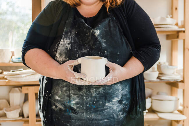 Cropped unrecognizable adult female artisan in dirty apron and black clothes standing in light studio and holding handmade ceramic pot near clayware — Fotografia de Stock