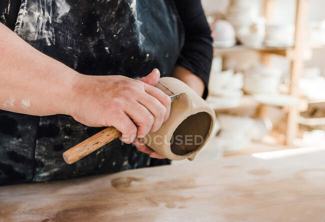 Неузнаваемая ремесленница в грязном черном фартуке и повседневной одежде, стоящая у стола и формирующая глиняную вазу профессиональным ножом в мастерской днем возле полок с глиняной посудой — стоковое фото