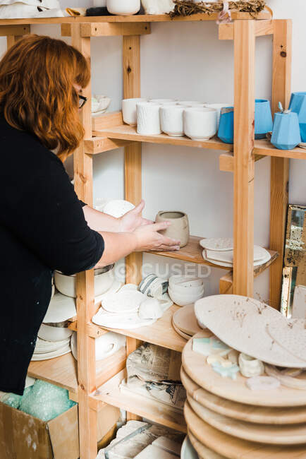 Side view of unrecognizable woman in casual black outfit standing near shelves with various pots and bowls with plates and different pottery while putting small clay vase in light studio - foto de stock