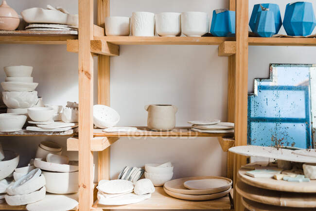 Collection of handmade ceramic bowls and vases with pots and plates with old mirror near different types of utensil standing on wooden shelves in light studio - foto de stock