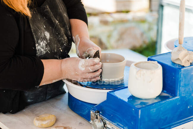 Анонимная женщина-ремесленница в фартуке моделирует глиняный горшок и корректирует форму на бросающем колесе — стоковое фото