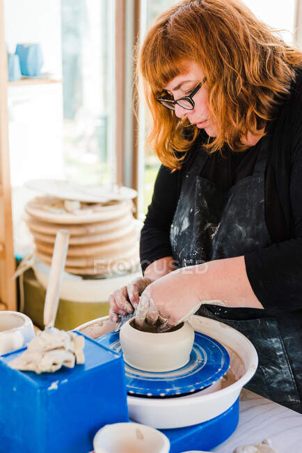 Сфокусированная женщина-ремесленница в фартуке моделирует глиняный горшок и корректирует форму на броске колеса — стоковое фото