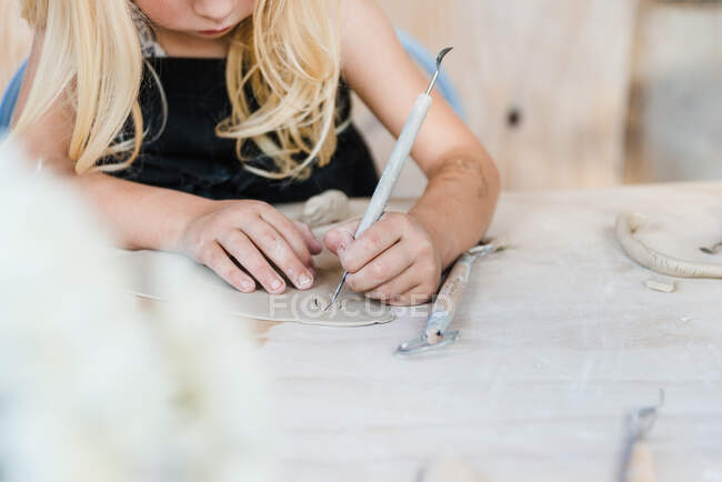 Crop anonymous little girl in black apron sitting at table and using double end loop while cutting piece of clay for pottery in light workshop — Stock Photo