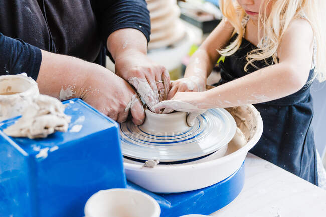 Crop unrecognizable female in black casual outfit standing near pottery wheel while shaping clay pot with little girl in light workshop — Stock Photo
