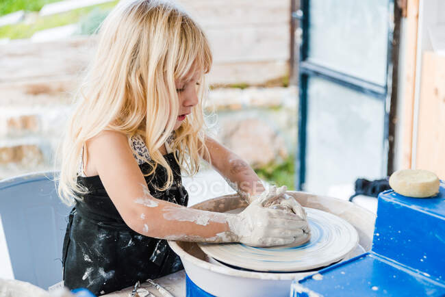 Вид сбоку на маленькую девочку в черном фартуке, стоящую рядом с гончарным кругом, формируя глиняный горшок в легкой мастерской — стоковое фото