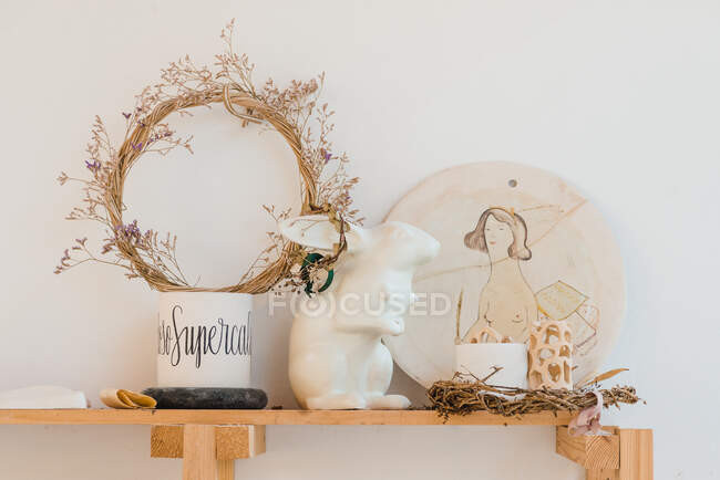 Couronne de plantes sèches près de la statuette de lapin et planche à découper peinte à la main près de tasse et objets décoratifs sur étagère en bois près du mur blanc — Photo de stock