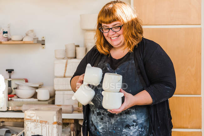 Lächelnde erwachsene Kunsthandwerkerin in schmutziger Schürze und schwarzer Kleidung, die im hellen Atelier steht und handgemachte Keramiktöpfe in der Nähe von Tonwaren trägt — Stockfoto