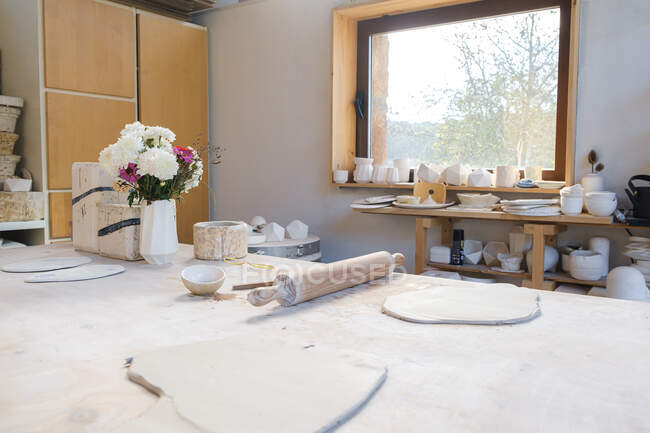 Table with smoothed pieces of clay near rolling pin and bowl near vase with flowers near window with  handmade pots on windowsill and plates on table in light studio - foto de stock