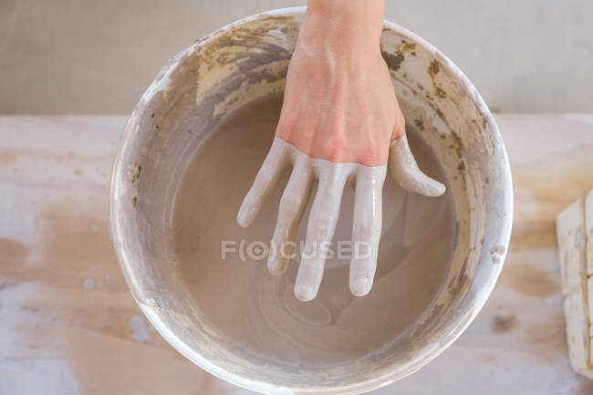 Anonyme Kunsthandwerkerin steht am Tisch und zieht im Atelier bei Tageslicht schmutzige Hand aus mit Ton gefülltem Eimer — Stockfoto