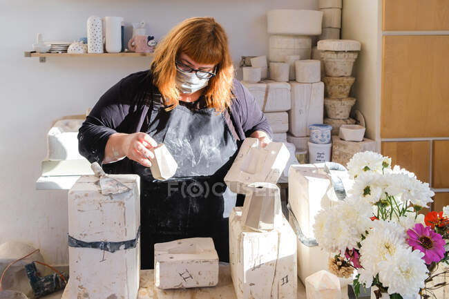 Female master in protective mask opening mild with handmade ceramic vase in pottery studio with blooming flowers — Foto stock