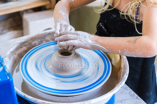 Crop unrecognizable little girl in black apron standing near pottery wheel while shaping clay pot in light workshop - foto de stock