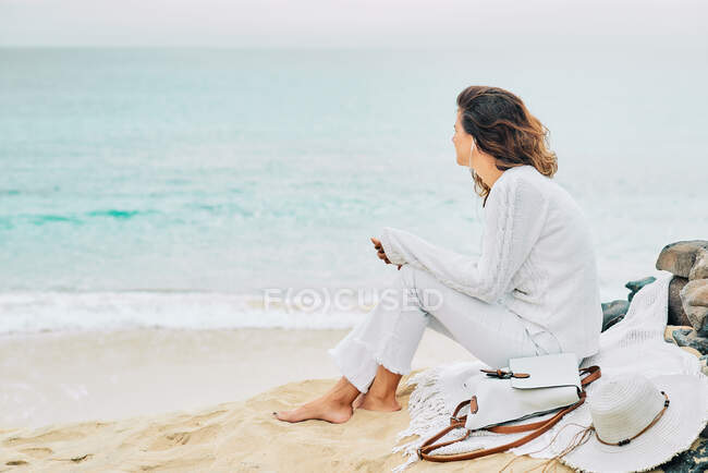 Side view of tranquil female sitting on beach near sea and enjoying music on earphones - foto de stock