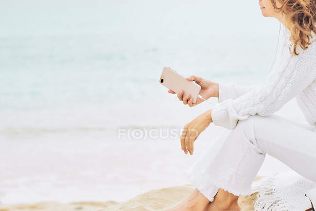 Side view of anonymous female sitting on beach near sea and enjoying music on earphones and using mobile - foto de stock