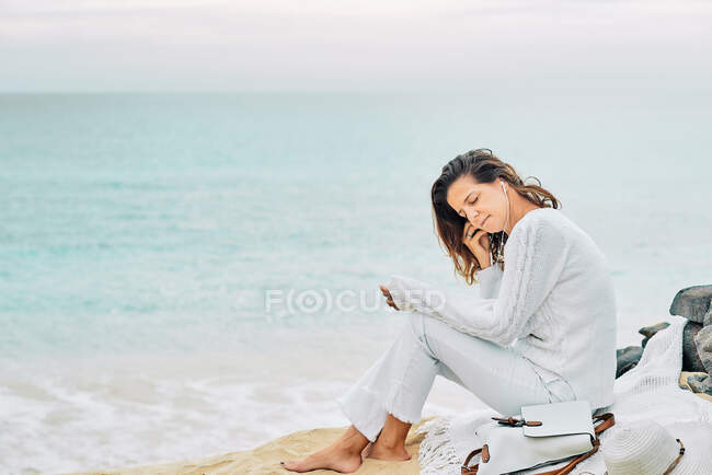 Side view of tranquil female sitting on beach near sea and enjoying music on earphones — Foto stock