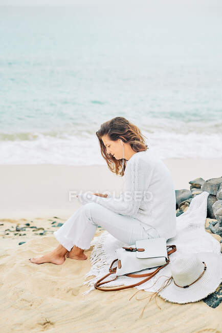 Side view of tranquil female sitting on beach near sea and enjoying music on earphones and using mobile - foto de stock